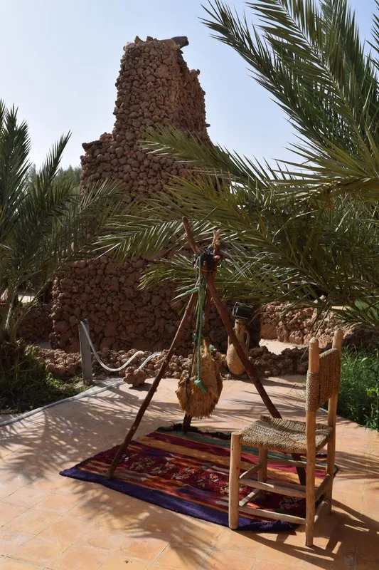 Les ruines d’une tour en terre parmi les palmiers ; au premier plan, un tapis et une chaise.