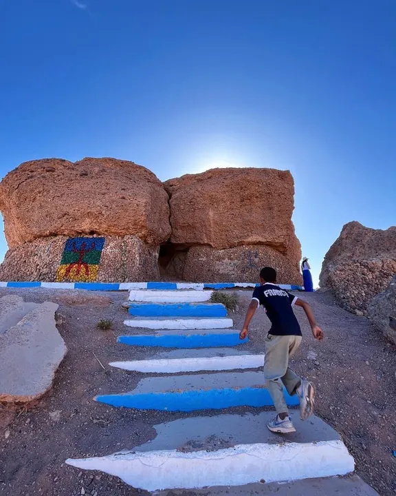 Un escalier peint en bleu et blanc grimpe entre des rochers vers le soleil ; un homme le gravit.