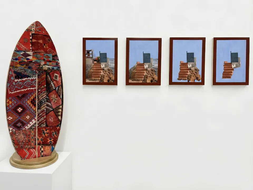 Vue d’installation : une planche de surf recouverte de motifs de tapis amazighs, à côté de quatre panneaux encadrés montrant un escalier et une porte bleue face au ciel.
